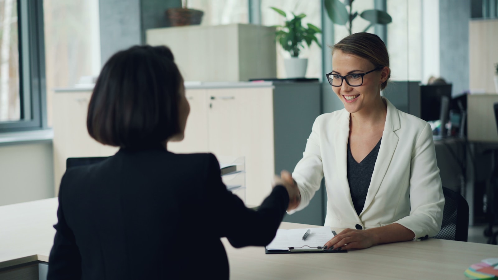 Two colleagues shaking hands over signed paperwork at an office desk